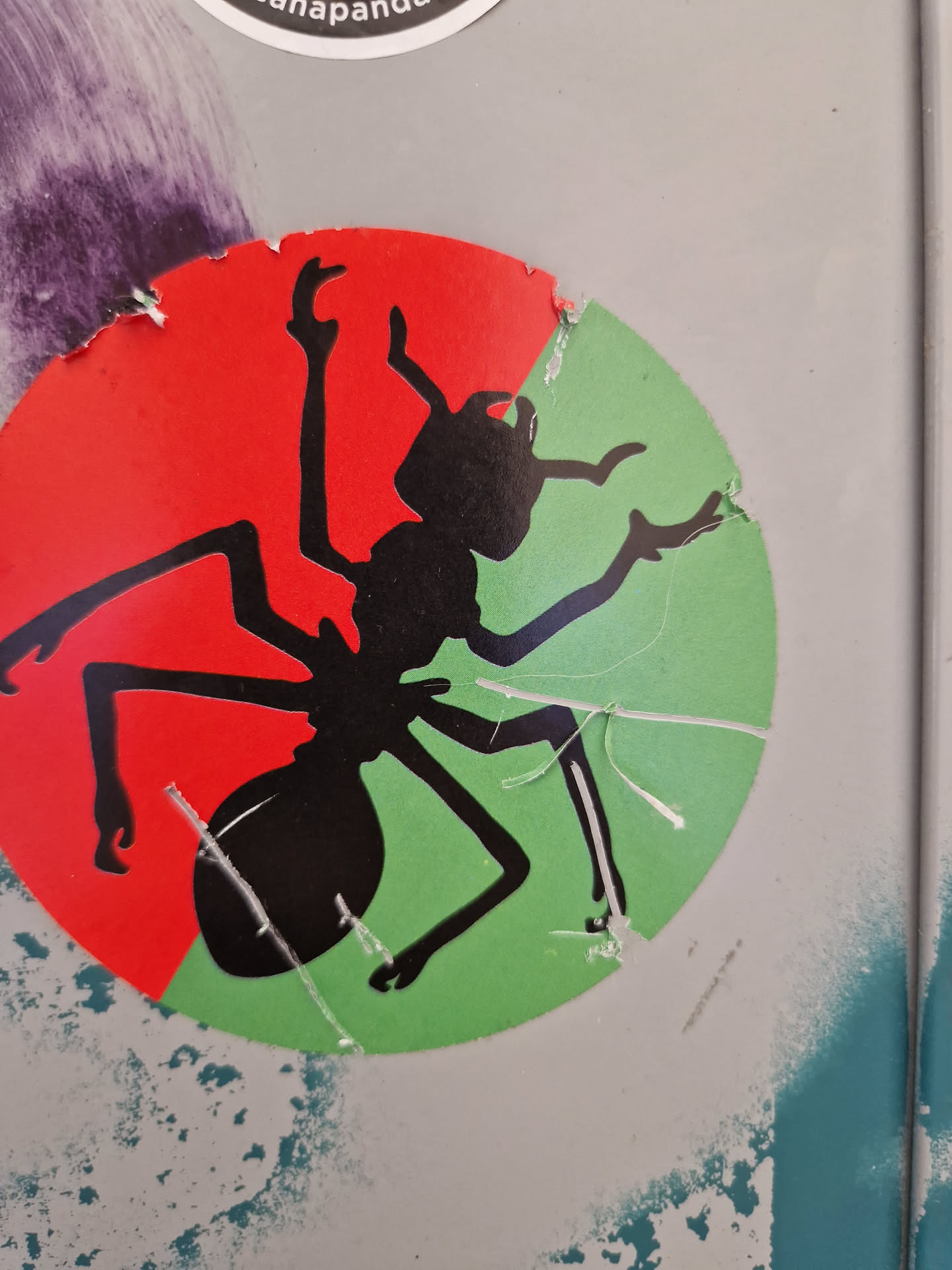 A round sticker featuring a black silhouette of an ant on a red and green background. The sticker shows some wear and tear from being placed outdoors.