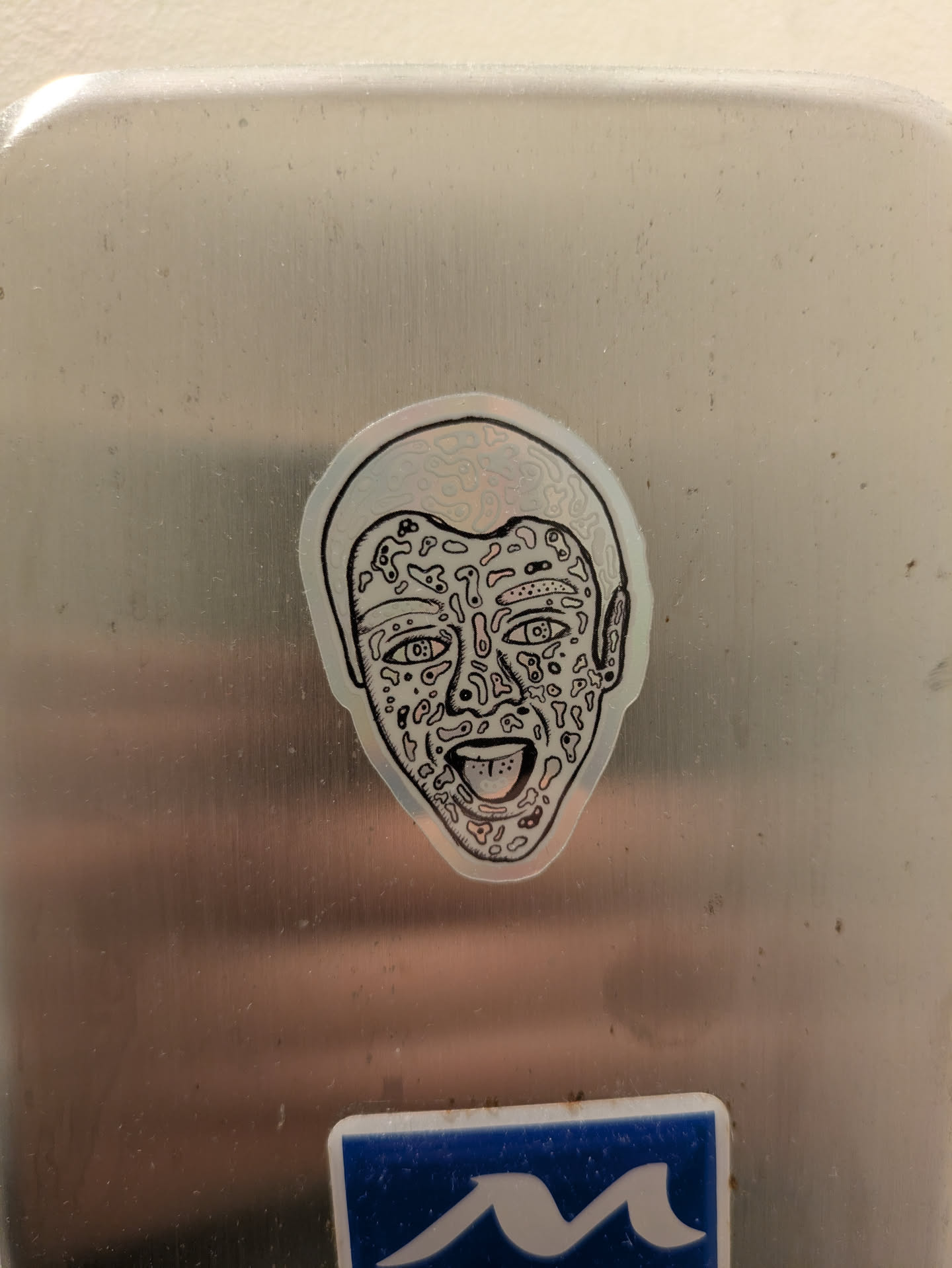 A silver sticker depicts a face with doodles and abstract patterns covering its surface. The face has its mouth open and tongue sticking out, giving it a goofy expression.