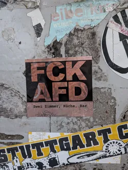 A square political sticker affixed to a weathered surface among other remnants of street postings. The sticker features a black central field with large bold letters spelling 'FCK' over 'AFD', patterned with a textured graphic in copper/rose gold tones. A band at the bottom displays the typewriter-style font text 'Zwei Zimmer, Küche, Bar' along with a faint stylized tag underneath.