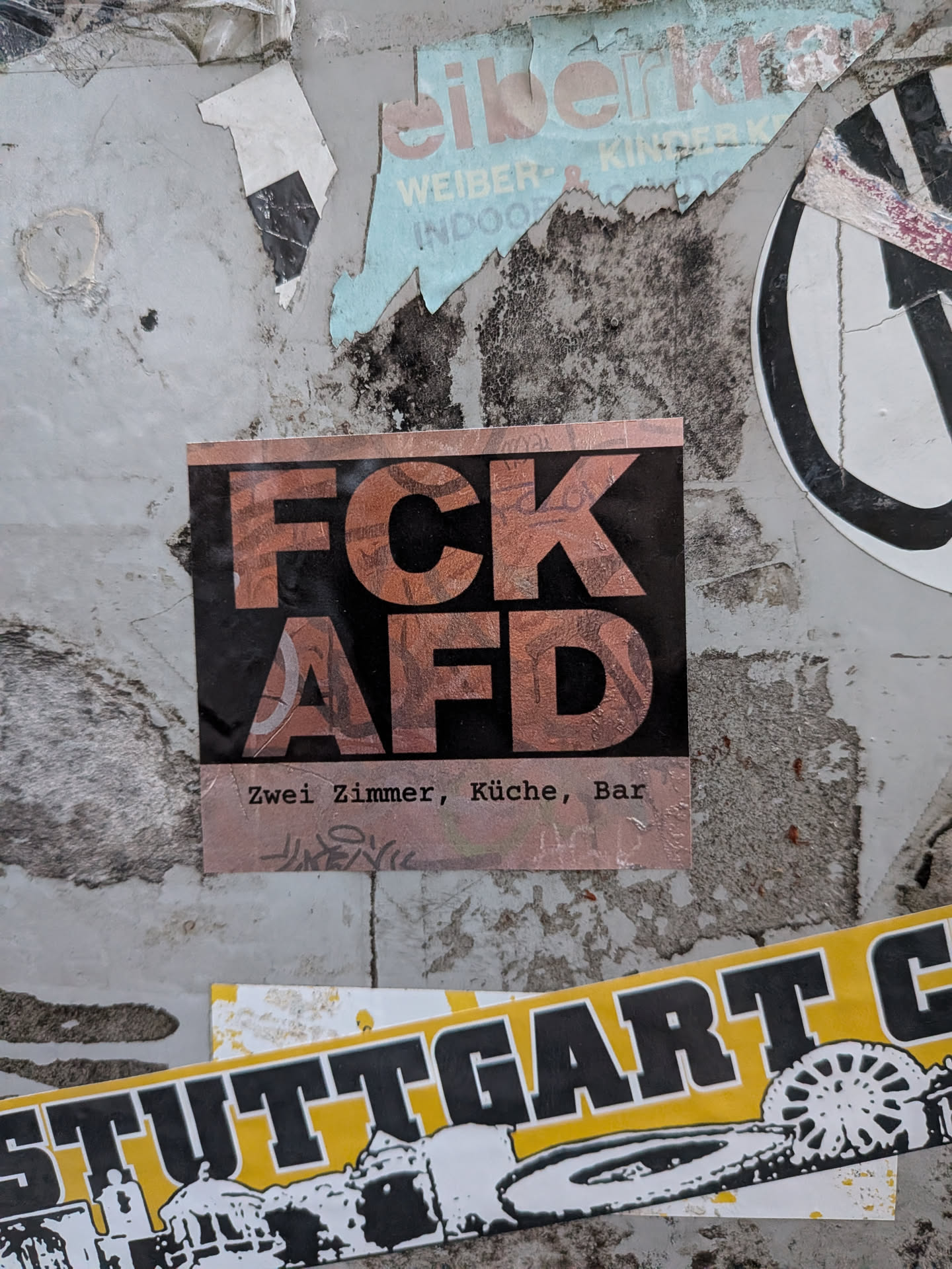 A square political sticker affixed to a weathered surface among other remnants of street postings. The sticker features a black central field with large bold letters spelling 'FCK' over 'AFD', patterned with a textured graphic in copper/rose gold tones. A band at the bottom displays the typewriter-style font text 'Zwei Zimmer, Küche, Bar' along with a faint stylized tag underneath.