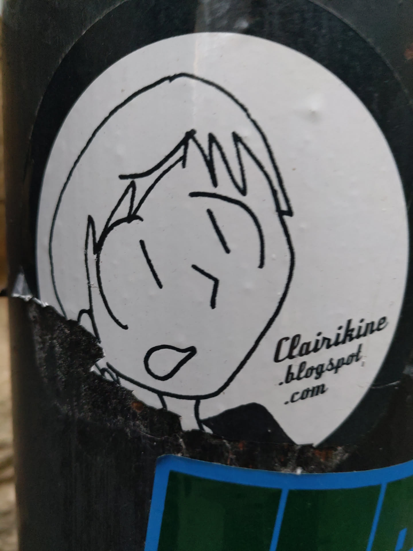 A white circular sticker with a simple drawing of a person's face with short hair. The drawing style is childlike. The text Clair ikine blogspot.com is visible at the bottom right corner of the sticker. 