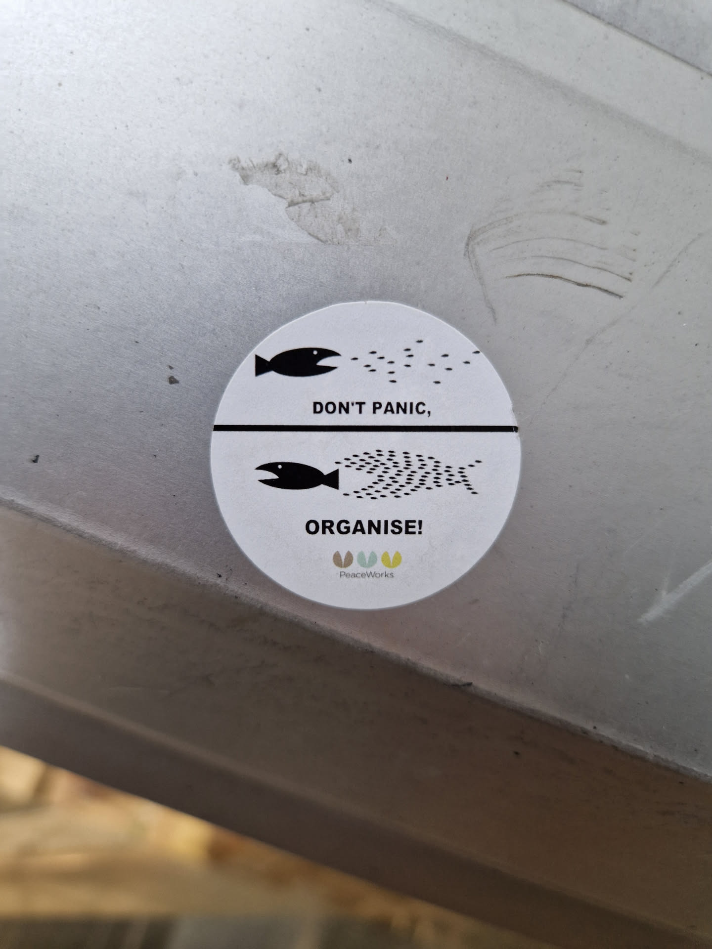 A round sticker with a minimalist design featuring two fish and the text 'Don't Panic, Organise!' It has a small logo at the bottom that says 'peaceworld'.