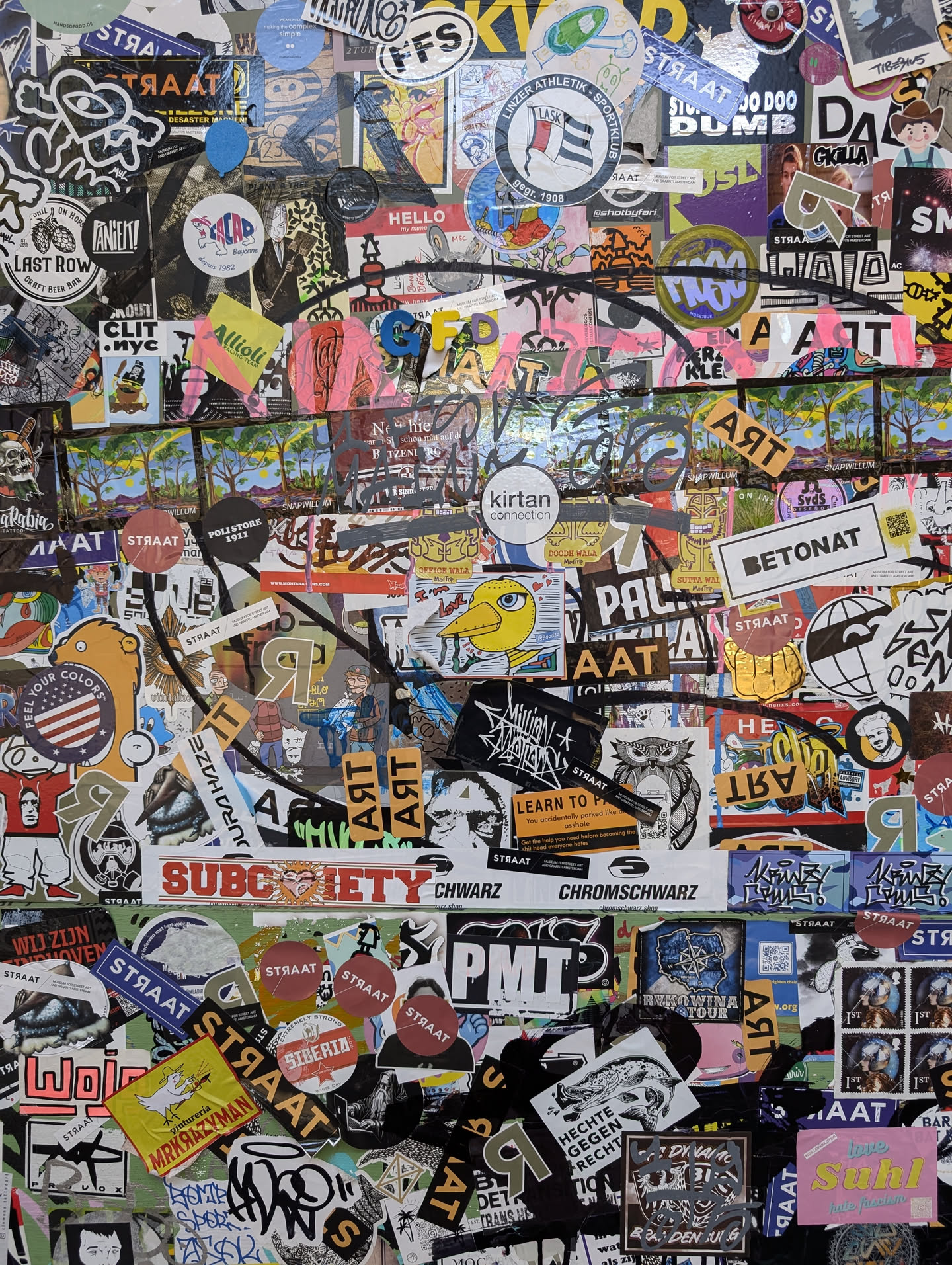 Close-up view of a wall covered in a multitude of stickers. The central focus is on a collection of stickers with various designs and text. Some prominent texts include 'Subciety', 'Chromschwarz', 'PMTP', 'Kirtan', 'Betonat', and 'Straat'.  Many stickers feature abstract designs, logos, and illustrations. The overall style is reminiscent of street art and sticker culture. No QR codes or social media links are visible.