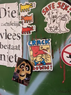 The sticker is white with a black outline and depicts a stylized, anthropomorphic condom. It has eyes, a mouth, and is pointing its finger upwards with a halo or sparkle above its head. In its right hand, it holds a rolled-up condom. The text 'SAFE SEX!' in red capital letters arches over the character. The background of the sticker is white, with some faint radiating lines behind the character.
