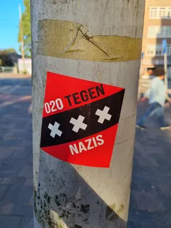 A red and black sticker with the text "020 TEGEN NAZIS" and three black crosses on a white background.  It is stuck on a grey pole.