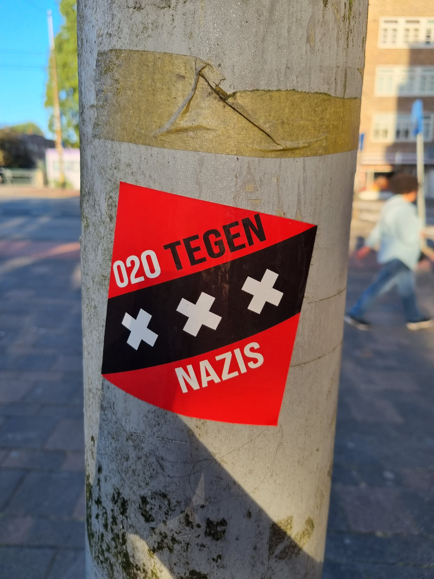 A red and black sticker with the text "020 TEGEN NAZIS" and three black crosses on a white background.  It is stuck on a grey pole.