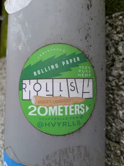 A green round sticker attached to a pole. The sticker advertises "ROLLS" rolling papers. It reads "UNBLEACHED ROLLING PAPER 100% PURE HEMP", with a drawing of a rolled cigarette and "WORLD'S LONGEST 20 METERS". It also features the website heavyrolls.com and the Instagram handle @HVYRLLS.