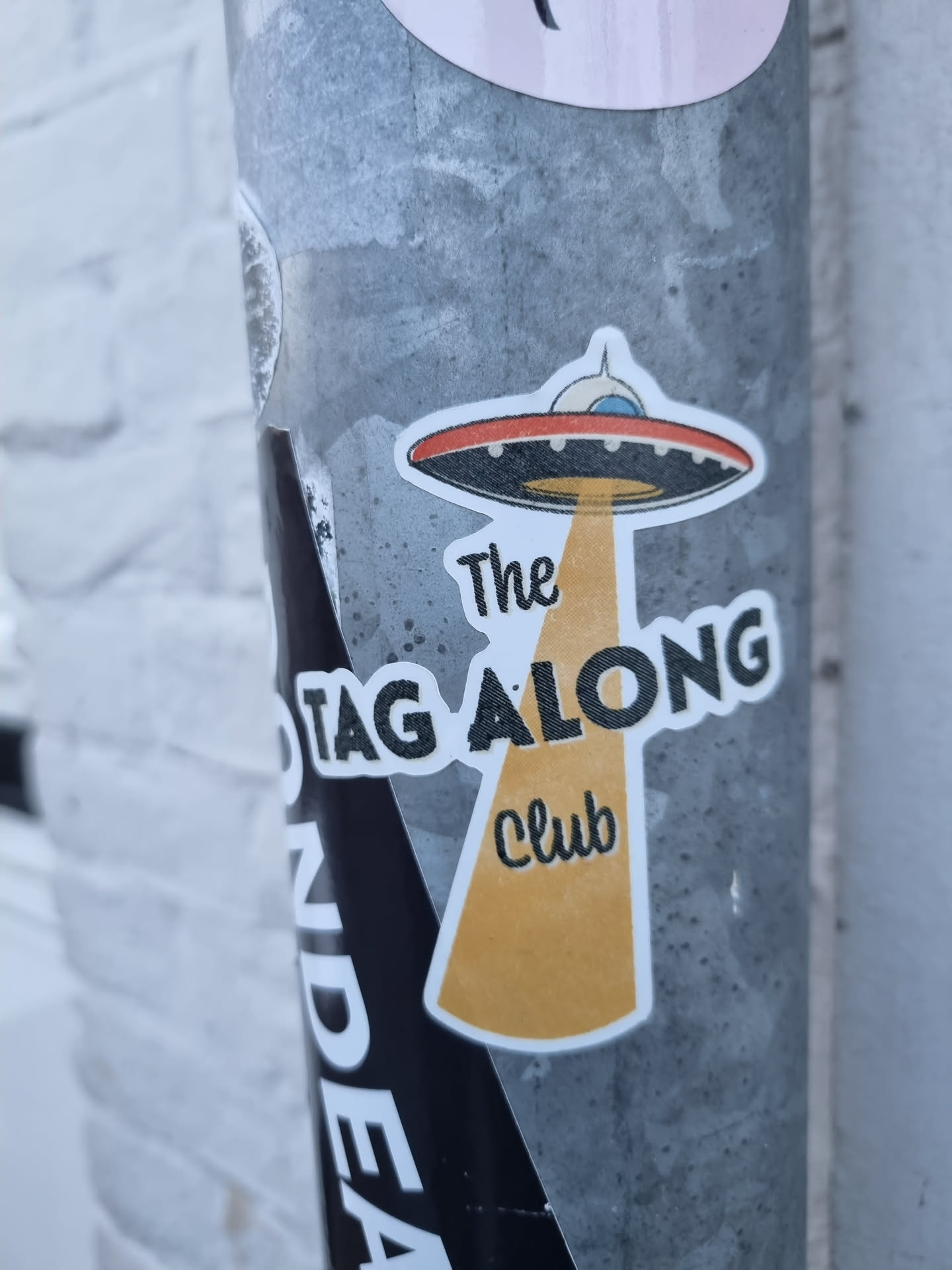 Sticker featuring an illustration of a UFO with a beam of light shining down. The text 'The Tag Along Club' is written below the UFO. 