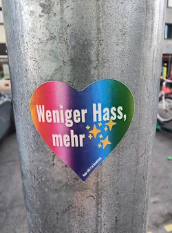 A heart-shaped sticker is affixed to a metal pole. The sticker features a rainbow gradient and the German text "Weniger Hass, mehr", which translates to "Less Hate, More". There are five yellow star designs adorning the sticker near the text. At the bottom corner, it says "Made with ❤️ in Osnabrück".