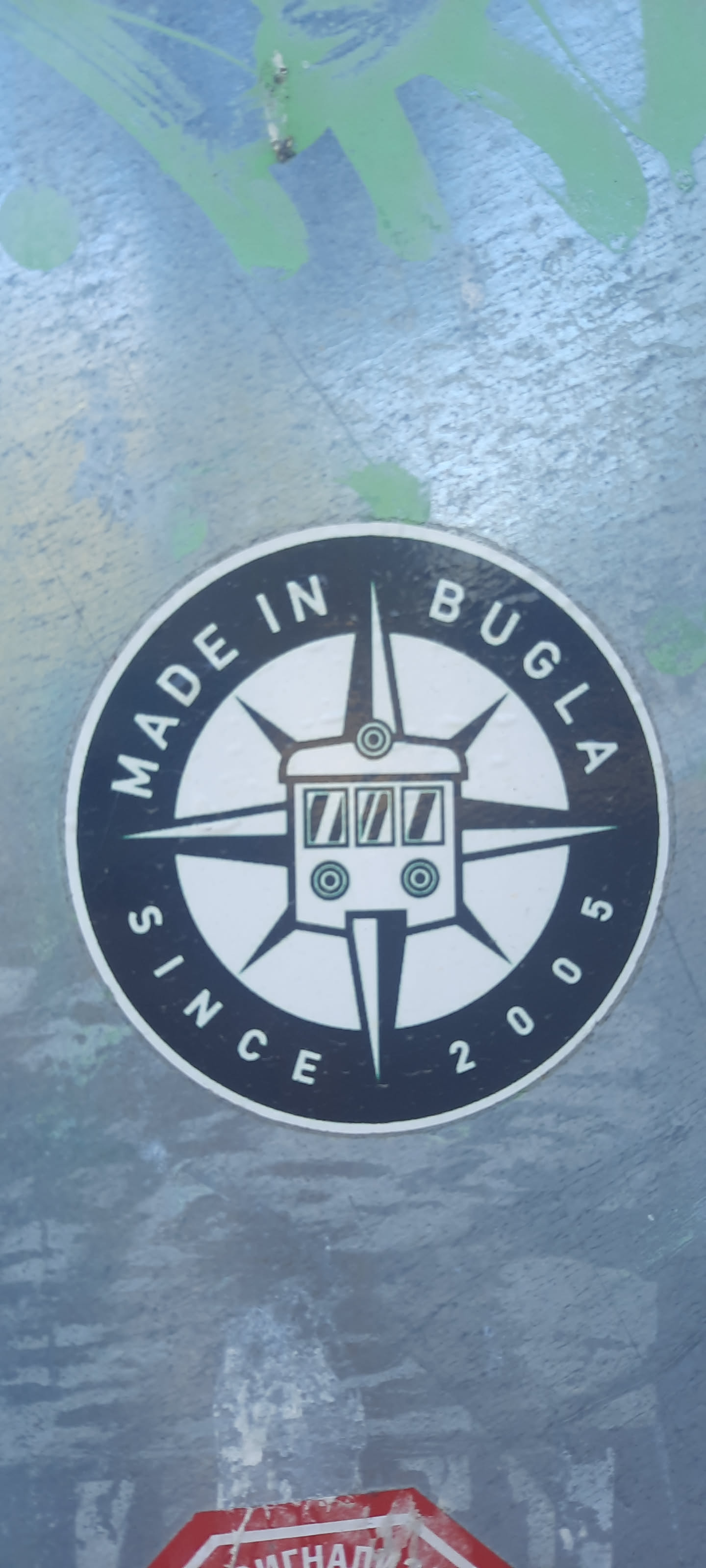 The sticker is circular, featuring a prominent black outer ring and a white inner circle. At its core is a stylized, front-facing depiction of a tram or bus, rendered in black outline with white fill, accented by small green details for its windows and lights. Behind the vehicle, a bold compass rose design extends with sharp, pointed arms, also in black and white. The black outer ring contains white text: 'MADE IN BUGLA' is curved along the top, and 'SINCE 2005' is curved along the bottom. The sticker is affixed to a textured, possibly metallic, surface, with remnants of green spray paint visible around its edges.