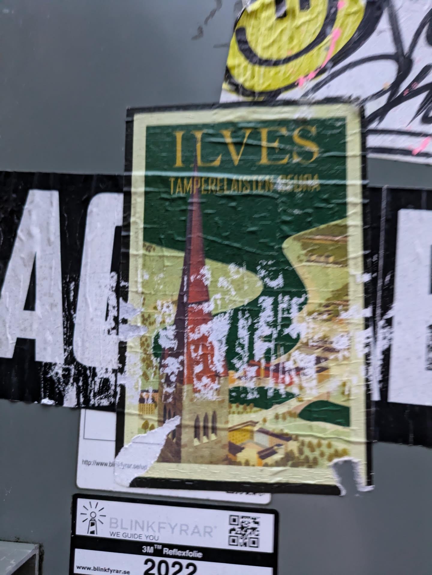 A vintage-style travel poster sticker featuring a picturesque illustration of a church and surrounding landscape, with the text "ILVES TAMPERE-LAISTEN SEURA" visible. The sticker shows some signs of wear and tear, suggesting it may have been affixed outdoors for some time. At the bottom of the image there's a smaller sticker with a QR code and a website address.