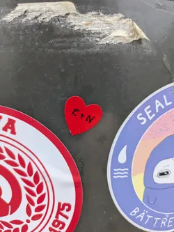 A red heart sticker with the letters E + N written in black marker. The sticker appears to be on a surface with other stickers.