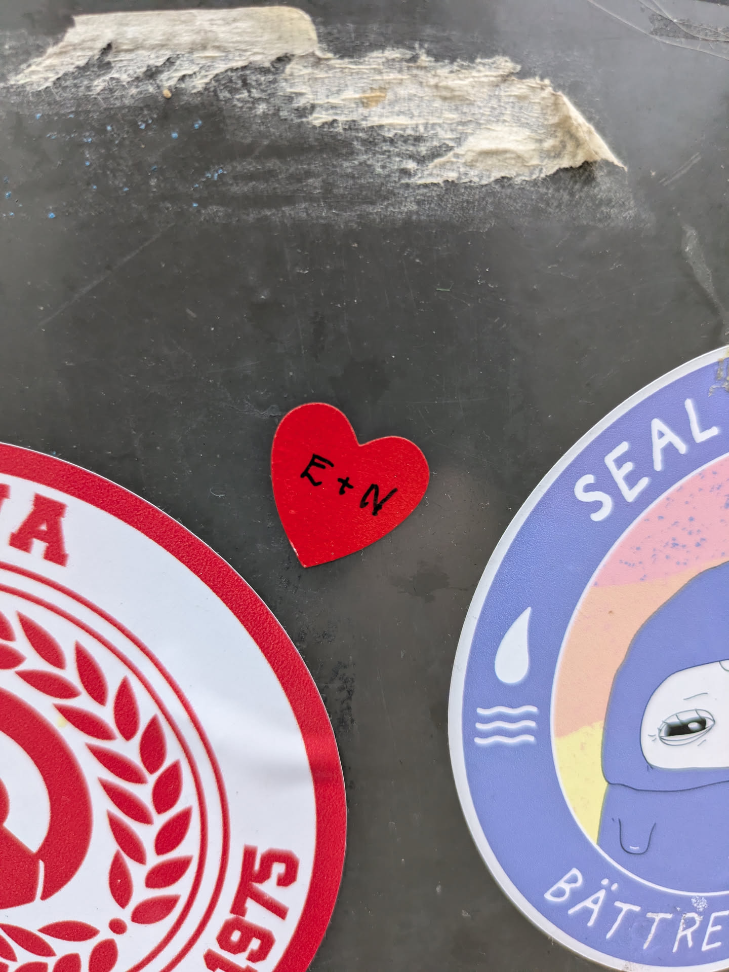 A red heart sticker with the letters E + N written in black marker. The sticker appears to be on a surface with other stickers.