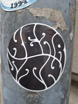 A round sticker with a dark background and a white design that looks like abstract art or a stylized plant.  The design is intricate and detailed. The sticker is affixed to a weathered surface.