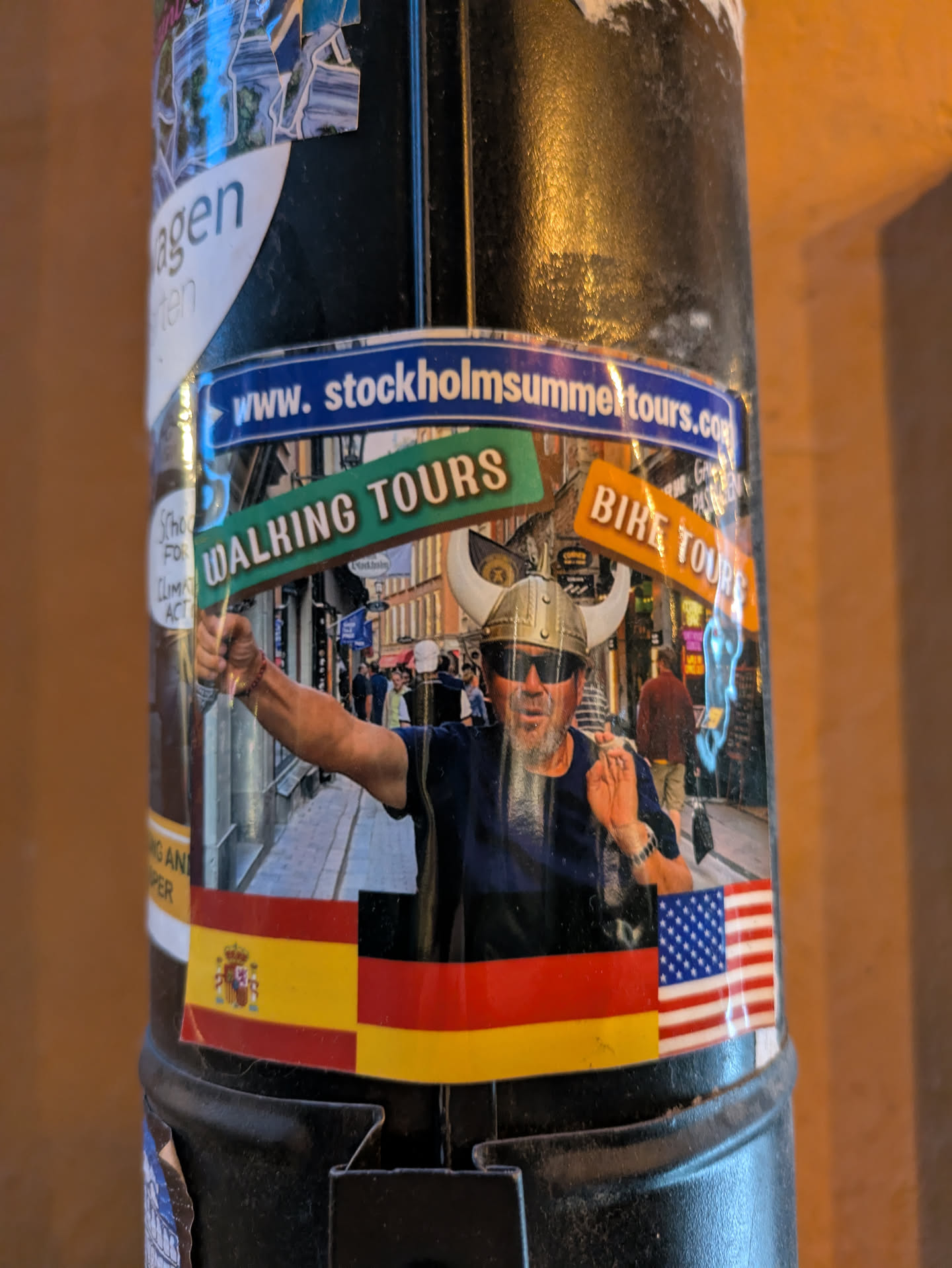 The central sticker features a man with a beard and a Viking helmet, wearing sunglasses and a blue shirt, with his arm outstretched. He is standing in a street scene that appears to be in Europe, with buildings and people in the background. Above him, there are green and orange signs that read 'WALKING TOURS' and 'BIKE TOURS' respectively. A blue banner above these signs reads 'www.stockholmsummertours.com'. At the bottom of the sticker, there are flags of Spain, Germany, and the USA. The sticker is slightly wrinkled and appears to be attached to a cylindrical object.
