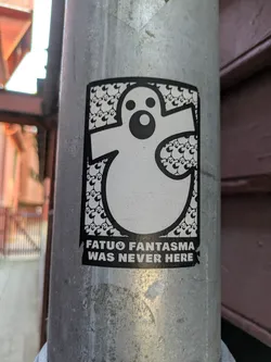 This sticker showcases a prominent black-outlined white ghost figure with simple, circular eyes and mouth. The interior of the large ghost is intricately filled with a repeating pattern of much smaller, identical ghost characters, creating a unique texture. Below the main graphic, white capitalized text on a black background reads 'FATUO FANTASMA' followed by a small ghost icon, and then 'WAS NEVER HERE'. The overall design is monochrome and features a playful yet slightly mysterious aesthetic. The sticker is affixed to a metallic pole.