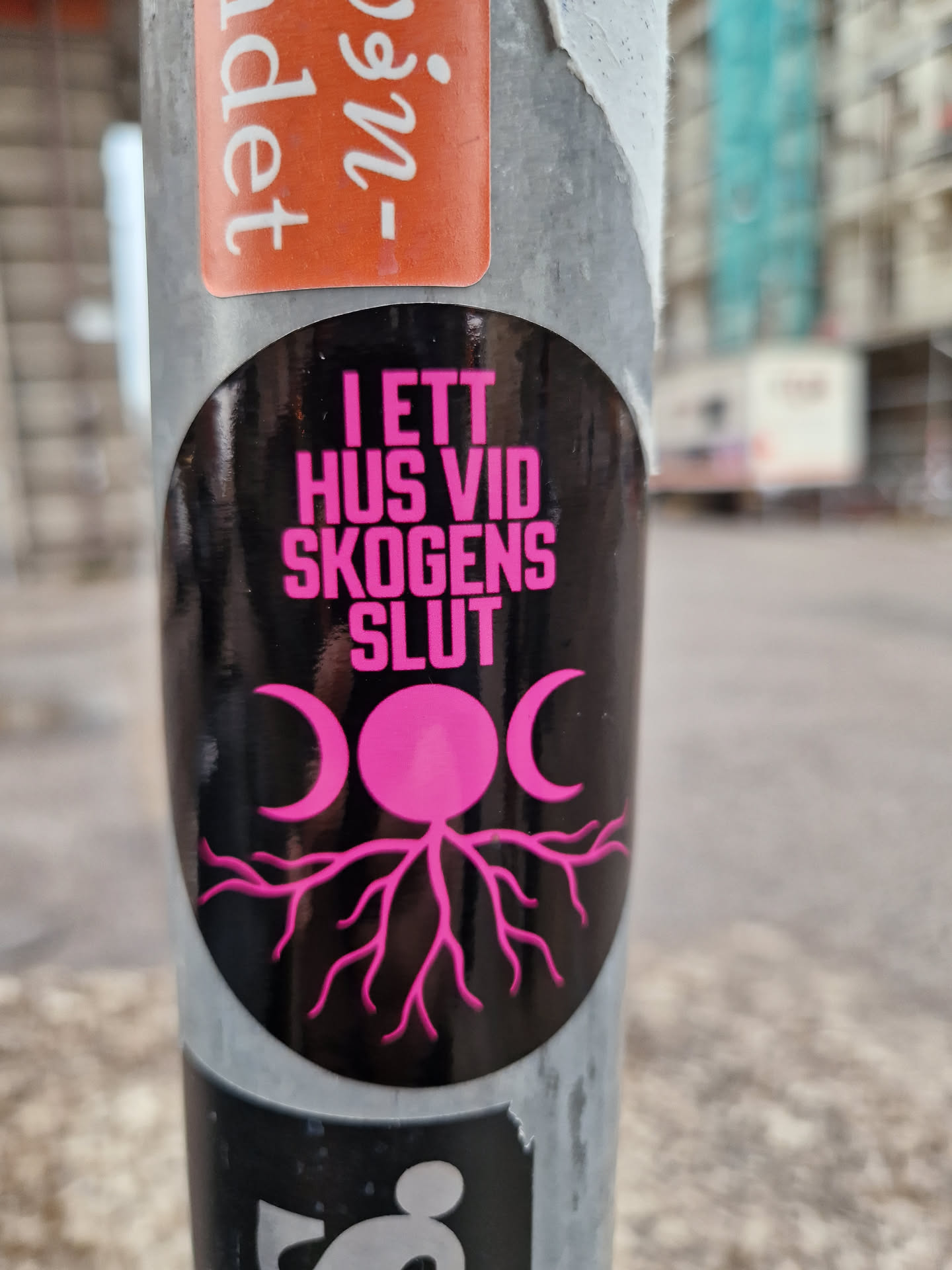 A black and pink sticker with text in Swedish that translates to "In a house at the edge of the forest." It features a pink moon phase symbol and tree roots design. The sticker is attached to a pole.