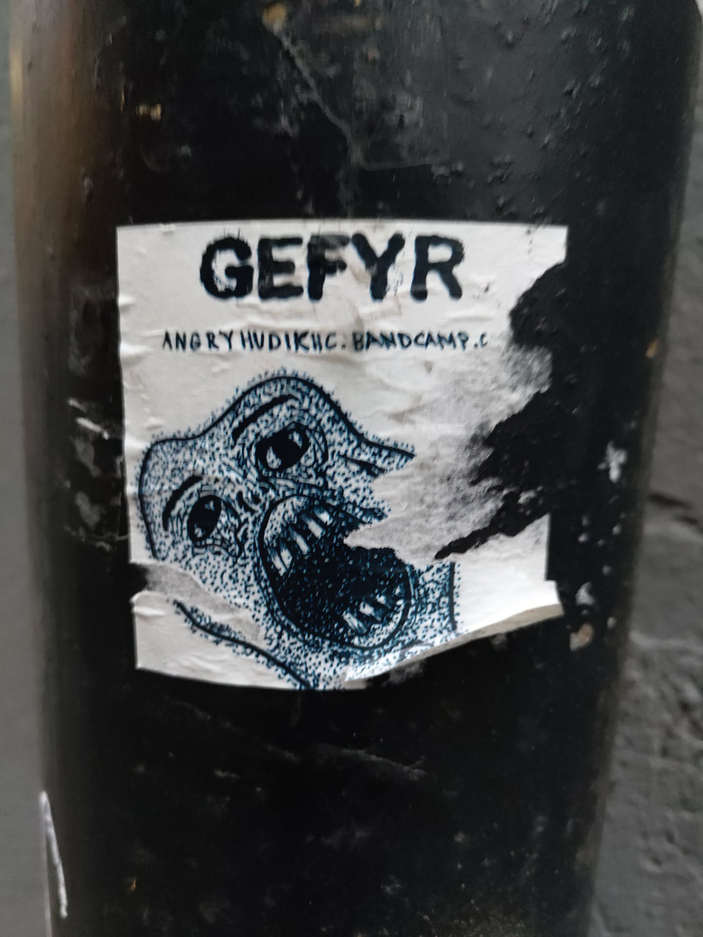 A sticker featuring the band name "GEFYR" and the text "ANGRY HUDIK HC BANDCAMP."  It includes a graphic illustration of a stylized creature with a large open mouth. The sticker appears to be affixed to a dark-colored surface, and part of it is torn or damaged.