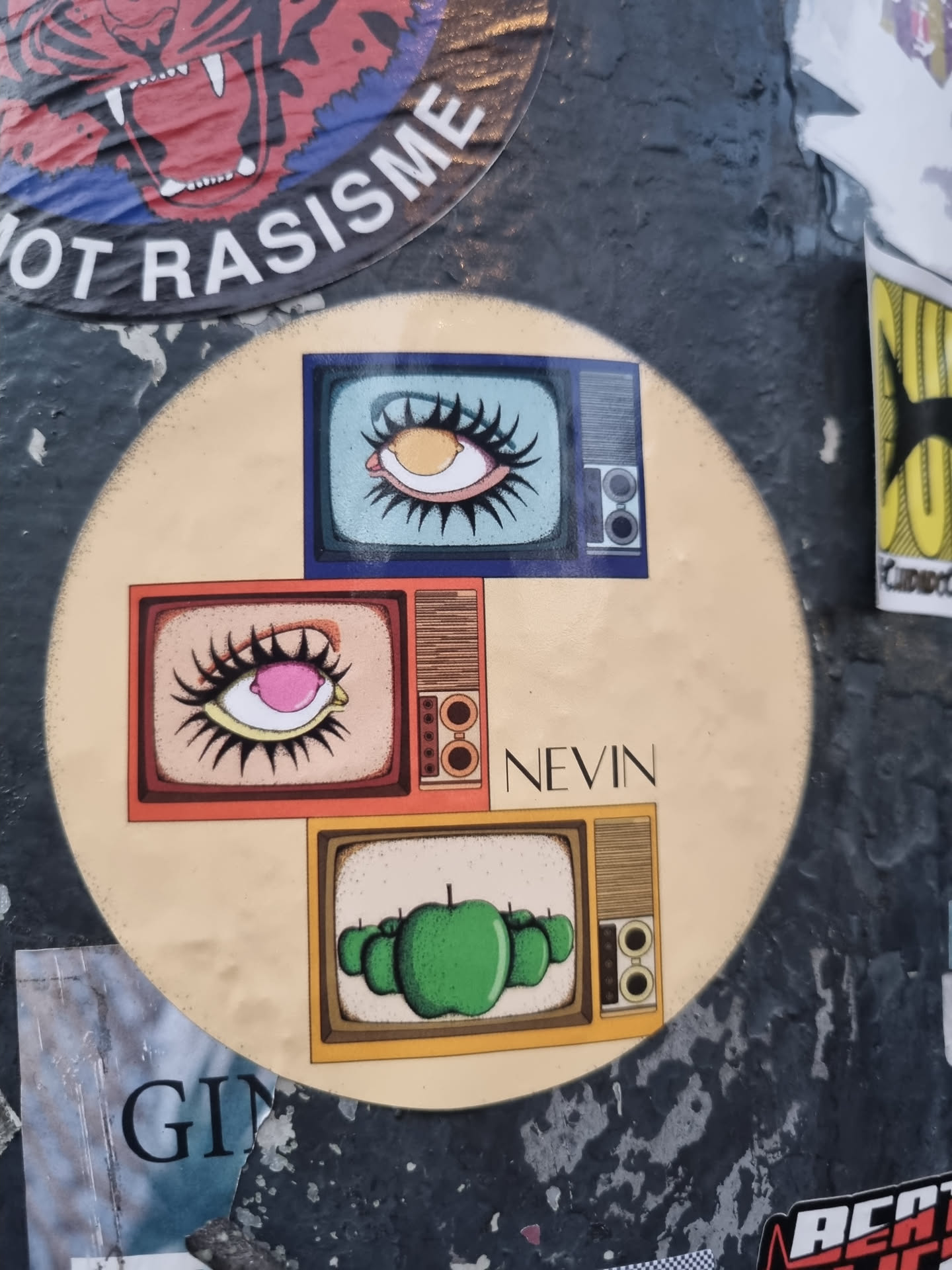 Round sticker featuring three retro TVs. Each TV displays a different image: a large eye with long lashes, another eye with a pink iris, and a group of green apples. The artist's name, Nevin, is written at the bottom. The sticker has a vintage, psychedelic aesthetic.