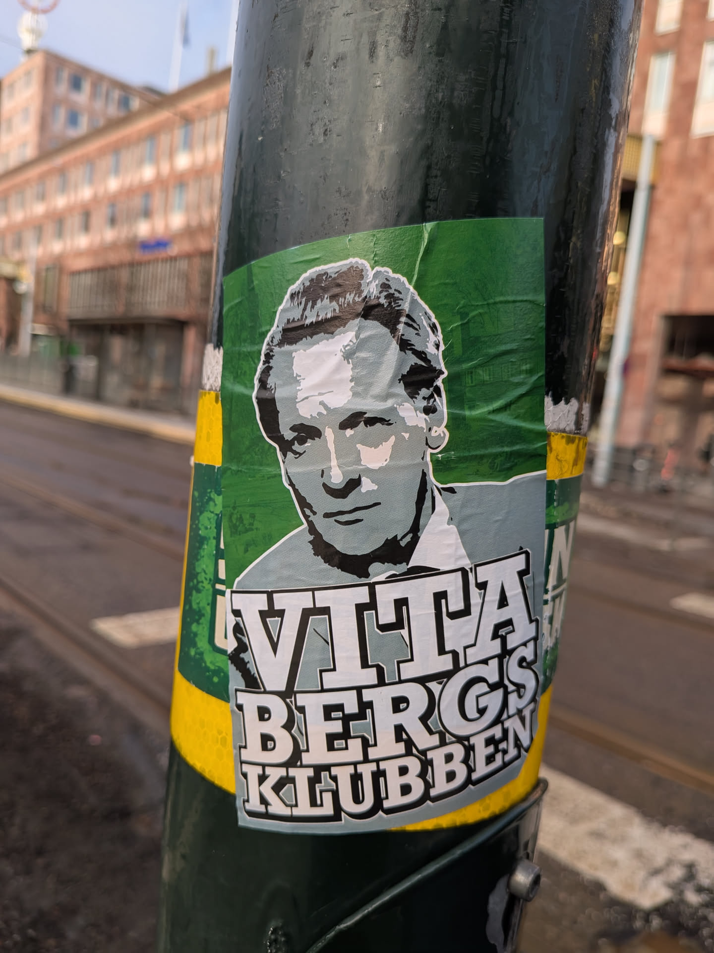 A rectangular sticker adhered to a dark green pole, featuring a stylized, monochromatic portrait of a man. The background of the sticker is a vibrant, somewhat crumpled green. The man in the portrait has short, wavy hair and a serious expression, rendered in shades of grey, black, and white, giving it a graphic, almost stencil-like appearance. Below the portrait, in large, bold, white block letters with a dark outline, is the text "VITABERGS" on the first line, and "KLUBBEN" on the second line. The sticker shows signs of wear, with slight wrinkles and peeling edges, particularly on the right side.