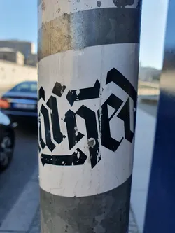 A sticker affixed to a metal pole features a stylized wordmark in black, set against a white background. The lettering is reminiscent of a gothic or blackletter typeface, with sharp angles and elongated forms.  The sticker shows some wear and tear, with minor scuffs and creases visible.