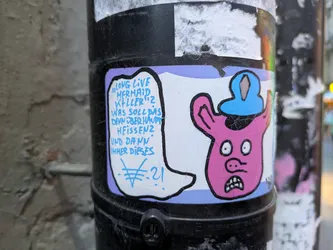 The sticker features a cartoonish pink creature with large, wide eyes, a prominent nose, and a gaping mouth with visible teeth. The creature wears a light blue hat with a darker blue rim. To the left of the creature, a speech bubble in white outlines with a light blue background contains blue text. The sticker has a purple border. The overall aesthetic is hand-drawn and somewhat crude, with visible cracks in the sticker material.