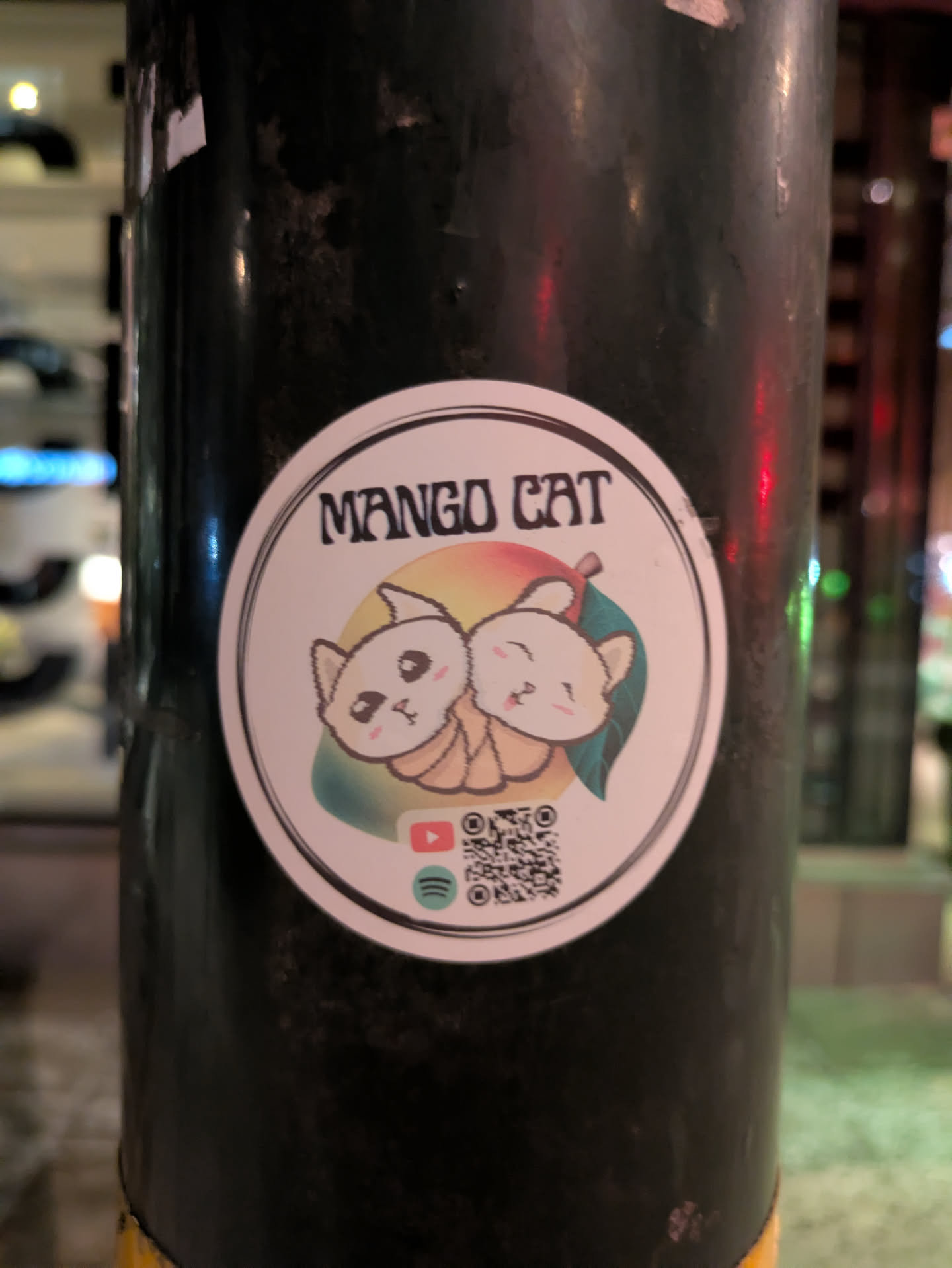 Sticker featuring two cute cartoon cats nestled together, resembling a mango. The sticker includes social media links to YouTube and Spotify, along with a QR code.