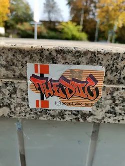 A close-up shot of a sticker featuring the word "TheDig" in stylized graffiti lettering. The sticker is orange and black and has a striped background. The Instagram handle "@board_doc_berlin" is visible at the bottom.