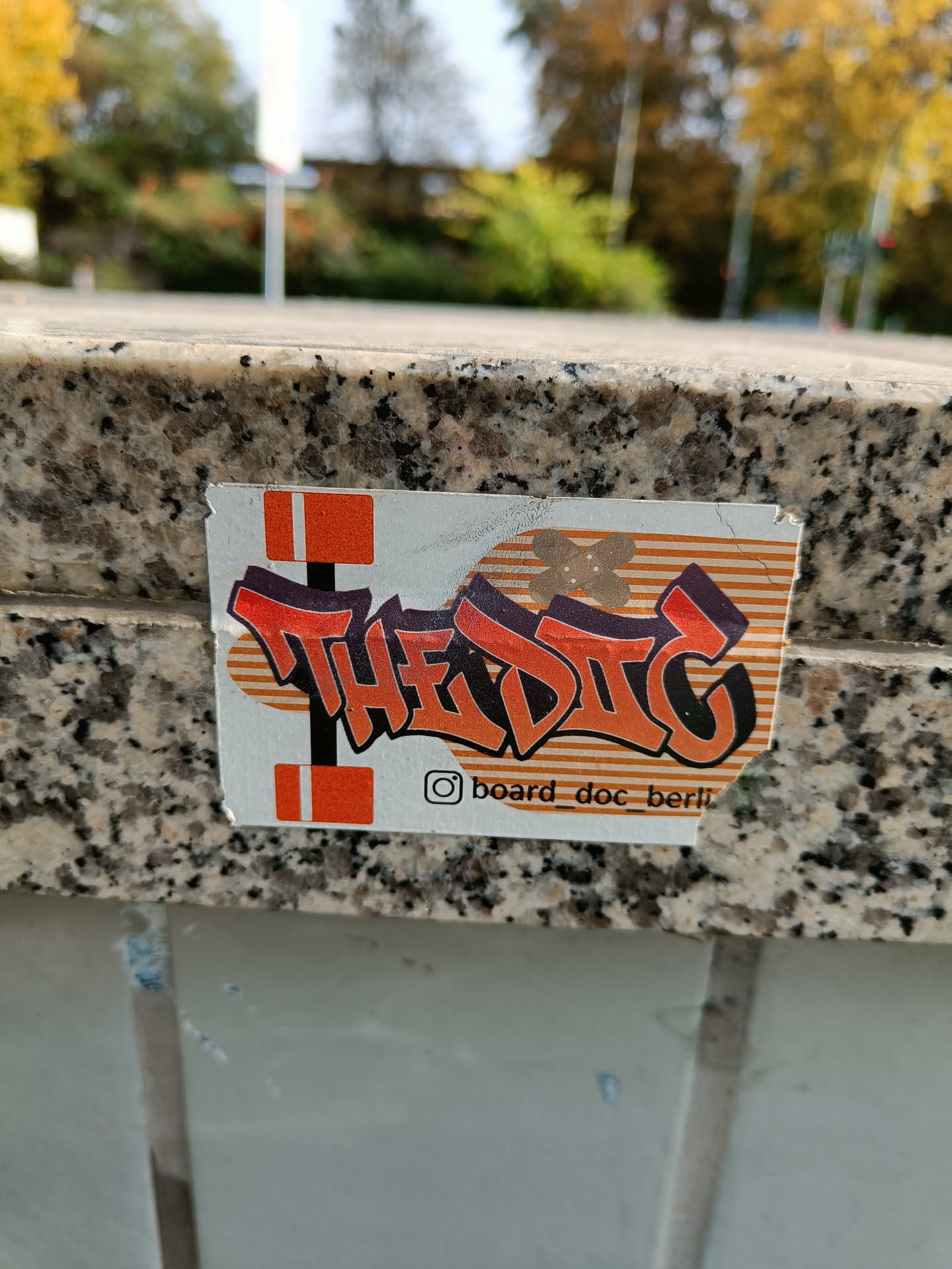A close-up shot of a sticker featuring the word "TheDig" in stylized graffiti lettering. The sticker is orange and black and has a striped background. The Instagram handle "@board_doc_berlin" is visible at the bottom.