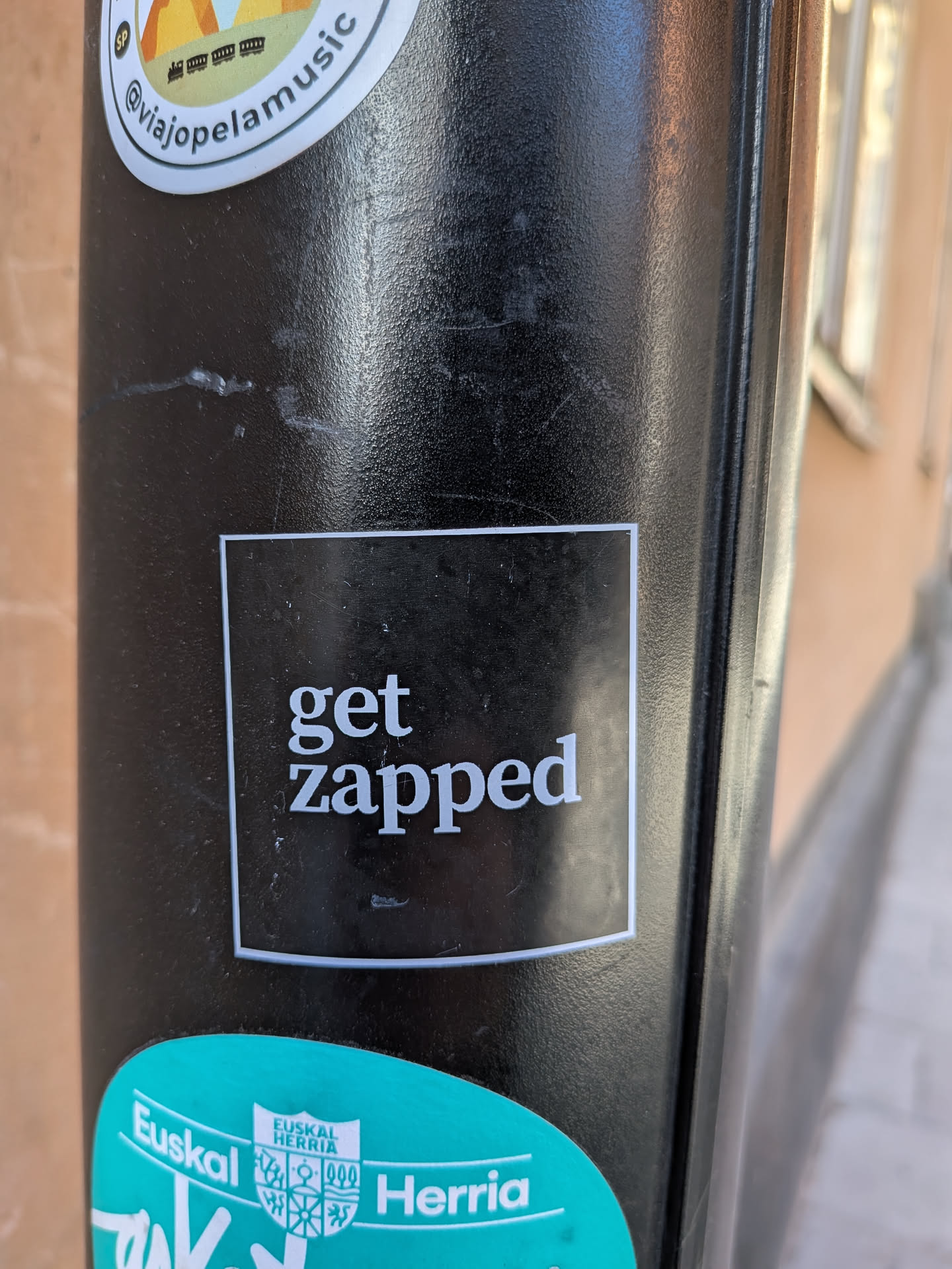 The sticker is a rectangular design featuring a stark black background framed by a thin white border. The phrase 'get zapped' is prominently displayed in white, classic serif typography. 'Get' is on the first line, and 'zapped' is on the second line, slightly larger, creating a clean and direct visual impact. The sticker appears to be affixed to a textured black pole.