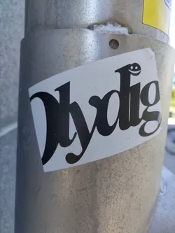Black and white sticker with the word "Hydig" written in a stylized, almost cursive font. A small smiley face is incorporated into the design. The sticker is affixed to a metallic pole.