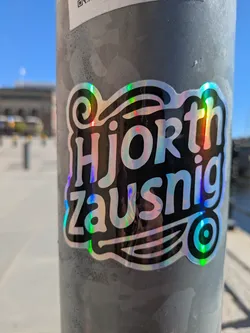 A glossy, holographic sticker of the words "Hjorth Zausnig" is stuck to a gray pole outdoors. The text is black with a swirly design above it, and a circular logo or drawing with concentric circles at the bottom right. The background reflects a building and blue sky. There is also another sticker above it.