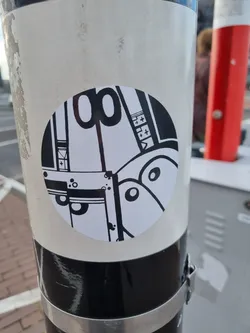 A round sticker with a black and white graphic design. The design appears to be abstract, possibly depicting a machine or mechanical object with various shapes and lines.  No visible text or QR code is present on the sticker itself.