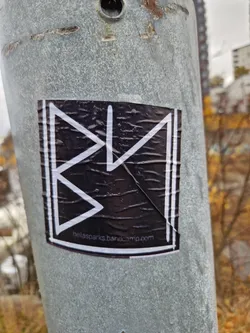 Black and white sticker with a geometric design and the text "bellaspks.bandcamp.com". The sticker is affixed to a metal pole outdoors.