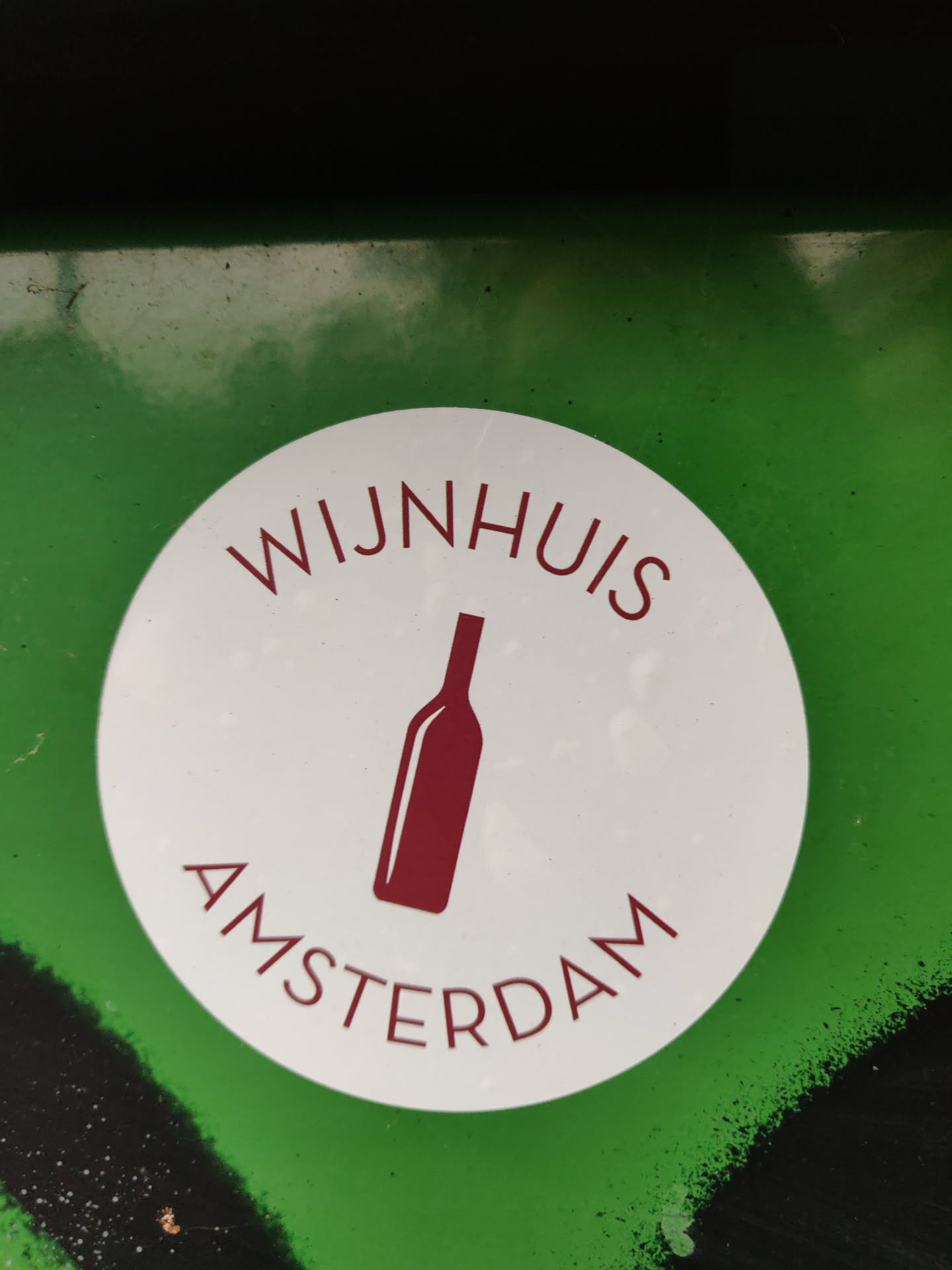 Round white sticker with a wine bottle icon and the text 'WIJNHUIS AMSTERDAM'. The sticker is on a green surface.