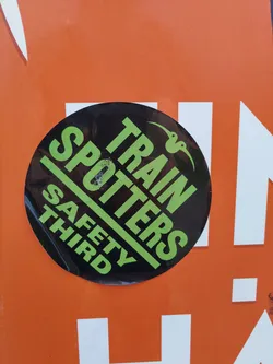 A round black sticker with the text "Train Spotters" in green and "Safety Third" in white. There is a small drawing of two birds in the upper right corner.