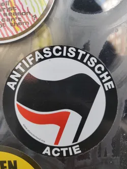 A round sticker with a black and red flag symbol and the text "ANTIFASCISTISCHE ACTIE" in a circle.  There is also a small website address at the bottom.