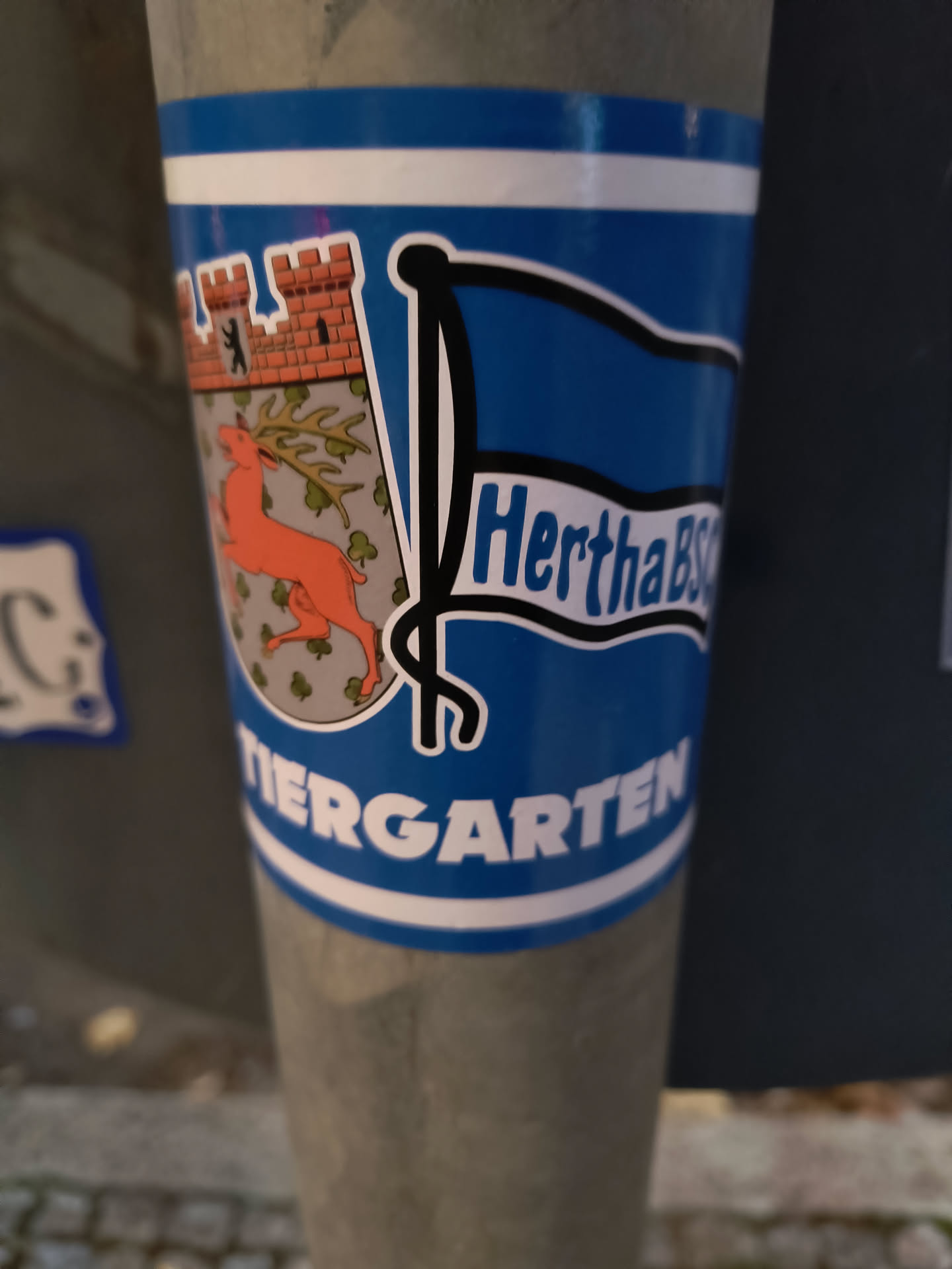 The sticker is predominantly blue and white, featuring a stylized flag design. On the left side of the flag, there is a coat of arms depicting a red deer with antlers, set against a background of green clovers and a stylized orange castle with a small black bear silhouette on its ramparts. The text 'HerthaBSC' is prominently displayed in white, outlined in black, on the blue part of the flag. Below the flag, the word 'TIERGARTEN' is written in large white capital letters, also outlined in black, on a blue background with white stripes above and below.