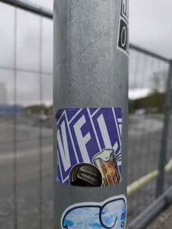 A square sticker on a metal post. The sticker has a purple background with "VFCL" written diagonally across it. There is a drawing of a soccer ball and a mug of beer in front of the text.