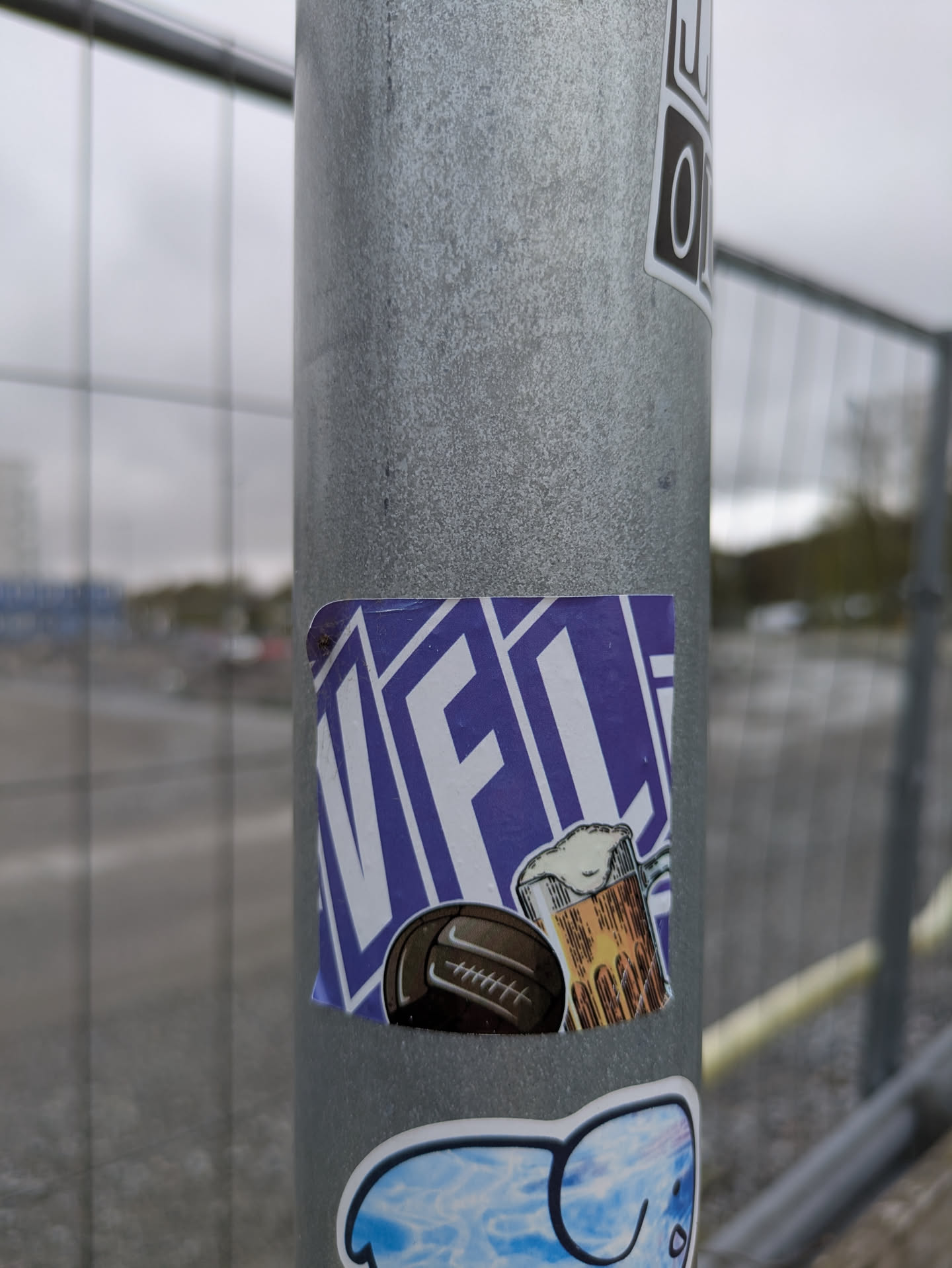 A square sticker on a metal post. The sticker has a purple background with "VFCL" written diagonally across it. There is a drawing of a soccer ball and a mug of beer in front of the text.