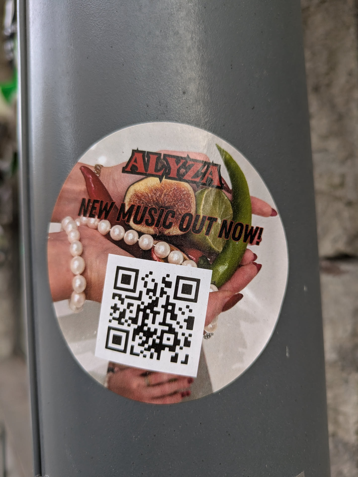 A circular sticker advertising new music by Alyza. The sticker features a hand holding fruits and peppers, adorned with a pearl necklace. A QR code is placed in the lower left.