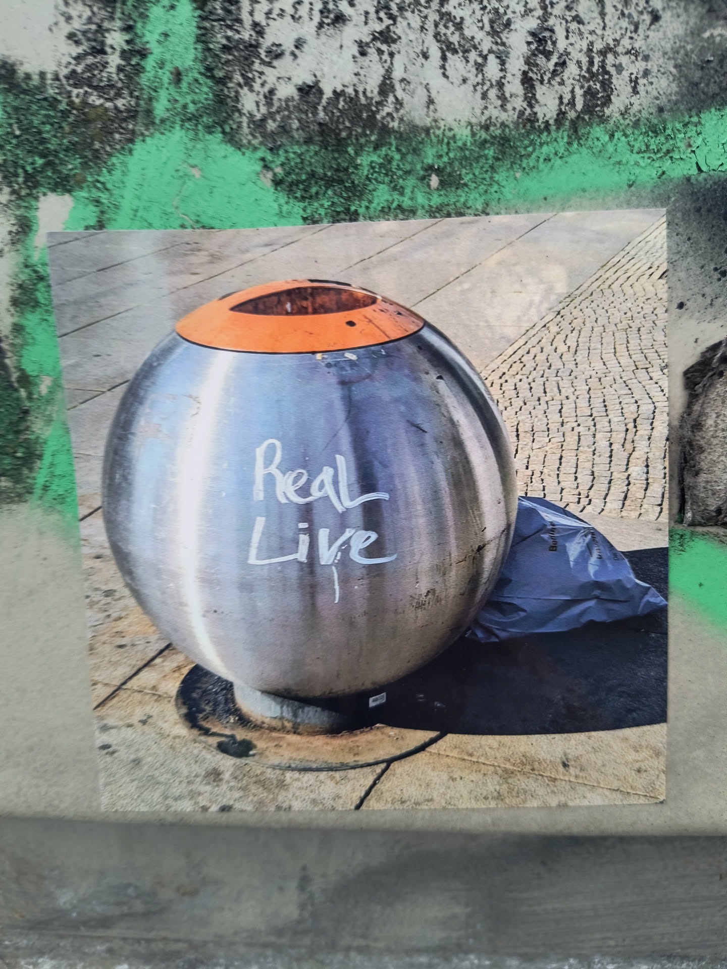 The sticker features a high-resolution photograph centered on a large, spherical, metallic silver waste bin with a contrasting orange rim and opening. White, handwritten text, reading 'Real Live,' is prominently visible on the bin's shiny surface. The waste bin stands on a paved ground, composed of both smooth concrete slabs and textured cobblestones. To the right of the bin, a dark, crumpled plastic bag rests on the ground. The lighting suggests an outdoor, daylight setting, emphasizing the metallic sheen of the bin. The sticker itself is a rectangular print, appearing slightly worn at the edges, adhered to a rough, painted wall.