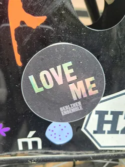 The sticker features a black background with the text 'LOVE ME' in a holographic, rainbow-reflective font. Below this, the words 'BERLINER ENSEMBLE' are printed in smaller font. The sticker is adhered to a black surface.