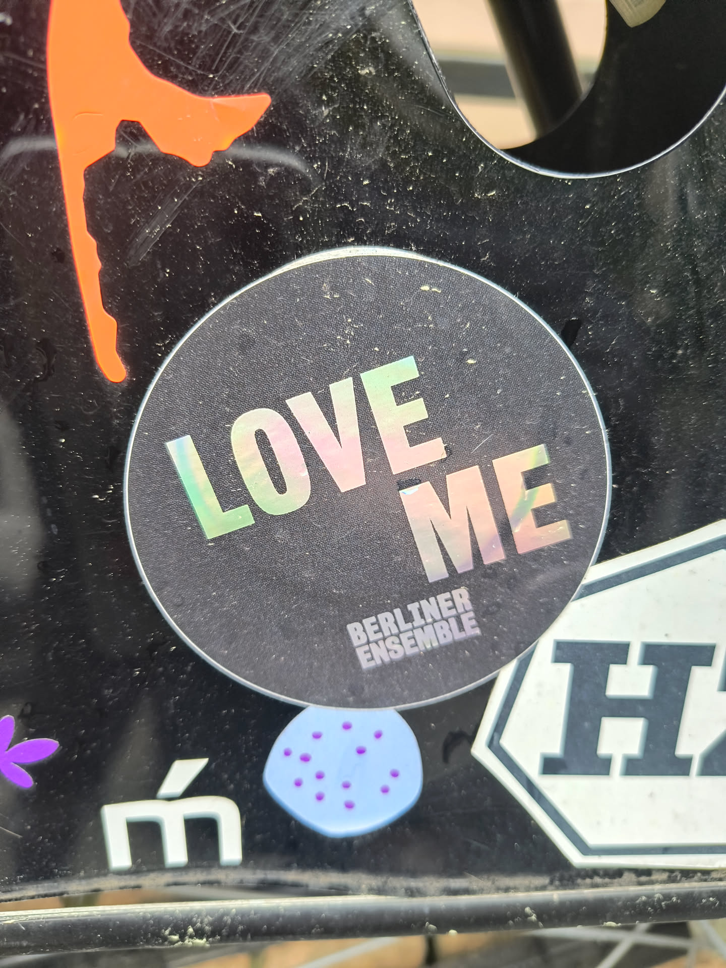 The sticker features a black background with the text 'LOVE ME' in a holographic, rainbow-reflective font. Below this, the words 'BERLINER ENSEMBLE' are printed in smaller font. The sticker is adhered to a black surface.