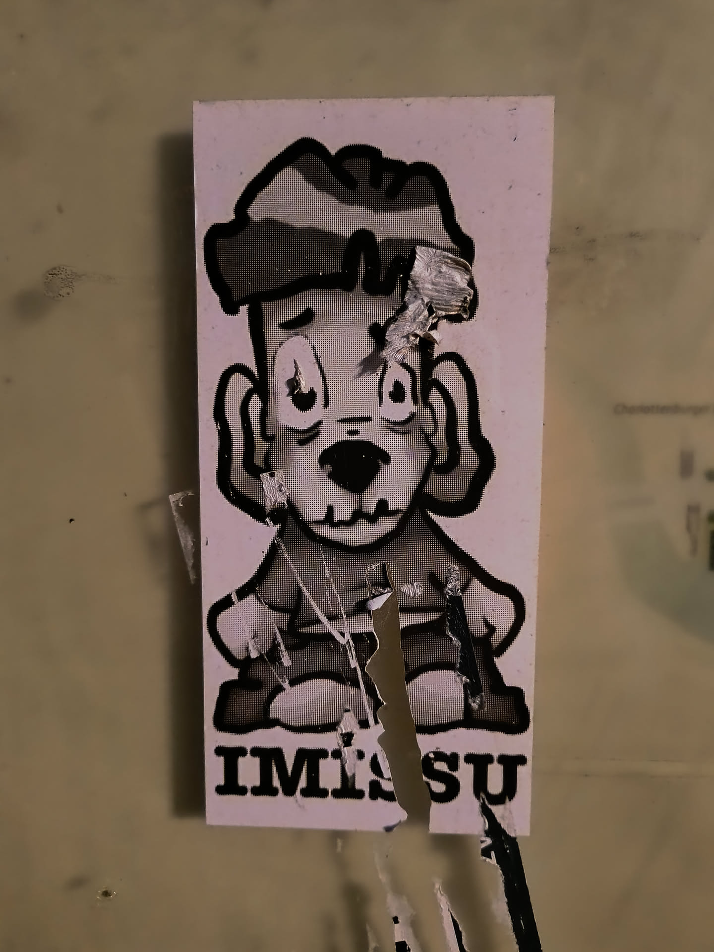 A sticker featuring a cartoon character with a dog-like face, wearing a hat and sitting down. The character is drawn in black and white with a somewhat distressed style, and the sticker itself shows signs of wear and tear. The text "IMISU" is printed below the character.