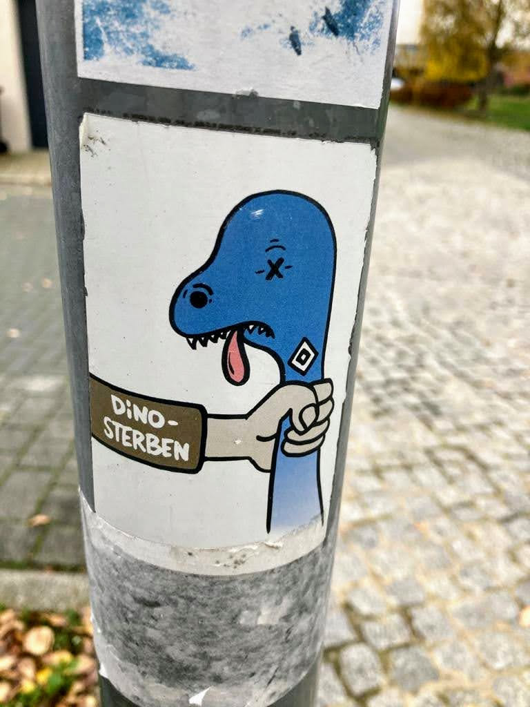 The sticker features a cartoonish blue dinosaur head with an 'X' for an eye and a drooping, pink tongue, indicating distress or death. Its neck is clutched by a pale-skinned hand, as if being strangled. On the wrist of the hand, a brown banner or sign is depicted, containing German text. A small, black and white diamond-shaped logo, resembling the Hamburger SV (HSV) football club emblem, is visible on the dinosaur's neck. The entire illustration is outlined in black on a white rectangular background, adhered to a grey pole, showing signs of wear and slight peeling at the edges.