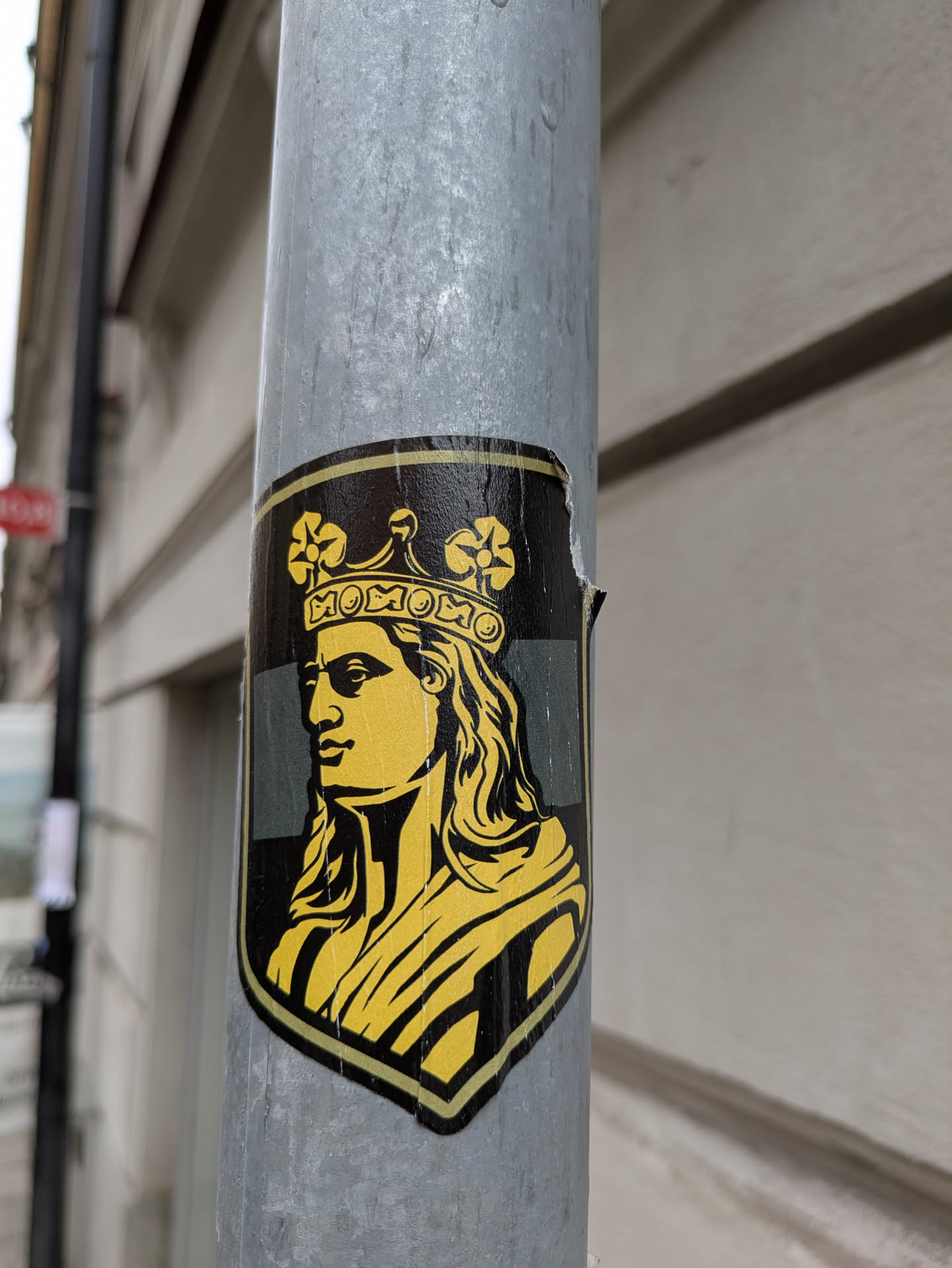 A black and yellow sticker depicting a stylized queen with the word "MOMOMO" above her head. The sticker has a shield shape and is stuck on a grey metal pole.