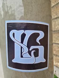 A square sticker with a light blue stylized letter G on a black background. The letter has decorative flourishes. The sticker is affixed to a grey surface.