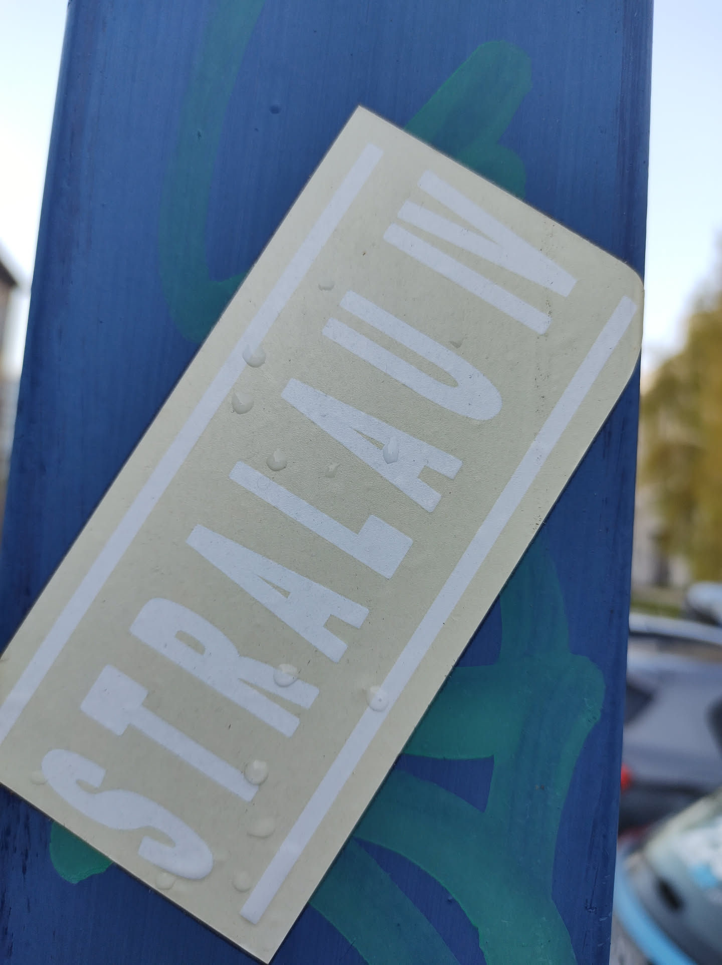 A beige rectangular sticker with the text STRALAU IV in white uppercase letters.  The sticker appears to be affixed to a blue surface outdoors and shows some water droplets.