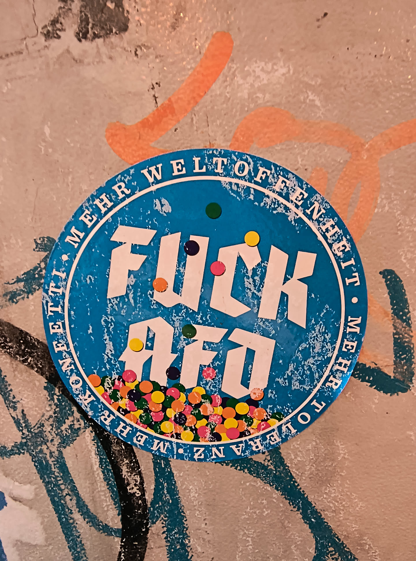 The sticker is circular and predominantly blue with white text and accents. The text 'FUCK AFO' is prominently displayed in large, bold white sans-serif font, with the letters 'FUCK' on the top line and 'AFO' on the bottom. Scattered around the text and within the bottom portion of the sticker are numerous small, colorful circles resembling confetti, in shades of pink, yellow, orange, green, and blue. The outer edge of the sticker features text in white, which appears to be German: 'MEHR WELTOFFENHEIT • MEHR TOLERANZ • MEHR KONEETTI'. The blue background has a distressed, textured appearance, with white speckles and scratches, suggesting wear and tear. The sticker is affixed to a wall with visible graffiti in the background.
